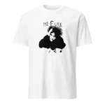 Uma mulher jovem e sorridente, com cabelo encaracolado, veste uma t-shirt branca de mangas curtas. A t-shirt tem uma estampa preta de Robert Smith, vocalista da banda The Cure. A mulher está a ajustar o cabelo com uma das mãos, enquanto a outra mão está na anca. O fundo desfocado mostra plantas de interior e móveis brancos, sugerindo um ambiente doméstico.