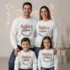 Uma família feliz (pai, mãe, filho e filha pequenos) a sorrir para a câmara, vestidos com sweatshirts brancas de Natal a condizer. As camisolas são personalizadas com o texto 'FAMÍLIA Silva' e 'CRIANDO MEMÓRIAS JUNTOS' em designs festivos com elementos natalícios como o Pai Natal, flocos de neve e árvores de Natal. O fundo é um cenário festivo e suavemente iluminado com luzes de fadas.