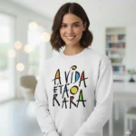 Uma mulher jovem e sorridente com cabelo castanho curto, a usar uma sweatshirt branca com a frase colorida 'A VIDA É TÃO RARA' estampada em estilo artístico. O fundo é um interior moderno e luminoso.