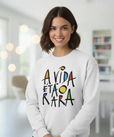 Uma mulher jovem e sorridente com cabelo castanho curto, a usar uma sweatshirt branca com a frase colorida 'A VIDA É TÃO RARA' estampada em estilo artístico. O fundo é um interior moderno e luminoso.