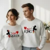"Casal jovem veste sweatshirts unissexo brancas a condizer. A sweatshirt da mulher tem a inscrição 'QUEEN' com a silhueta da Minnie a dar um beijo, e a do homem tem a inscrição 'KING' com a silhueta do Mickey. Entre ambos, pequenos corações vermelhos unem os desenhos."