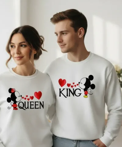 "Casal jovem veste sweatshirts unissexo brancas a condizer. A sweatshirt da mulher tem a inscrição 'QUEEN' com a silhueta da Minnie a dar um beijo, e a do homem tem a inscrição 'KING' com a silhueta do Mickey. Entre ambos, pequenos corações vermelhos unem os desenhos."
