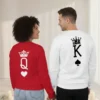 Fotografia de costas de um casal de mãos dadas, vestindo sweatshirts brancas unissexo. A peça da esquerda apresenta uma coroa vermelha sobre a letra Q e um coração; a da direita exibe uma coroa preta sobre a letra K e um símbolo de espadas.