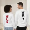 Fotografia de costas de um casal de mãos dadas, vestindo sweatshirts brancas unissexo. A peça da esquerda apresenta uma coroa vermelha sobre a letra Q e um coração; a da direita exibe uma coroa preta sobre a letra K e um símbolo de espadas.